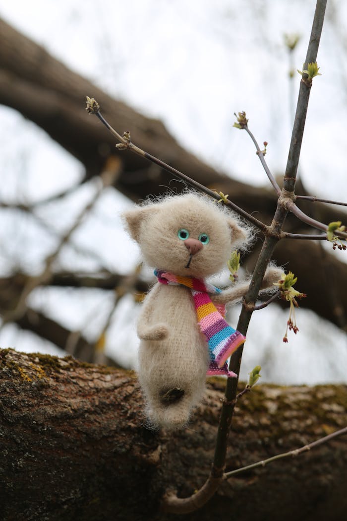 Teenused A whimsical felt cat toy wearing a colorful scarf perched on a budding tree branch.